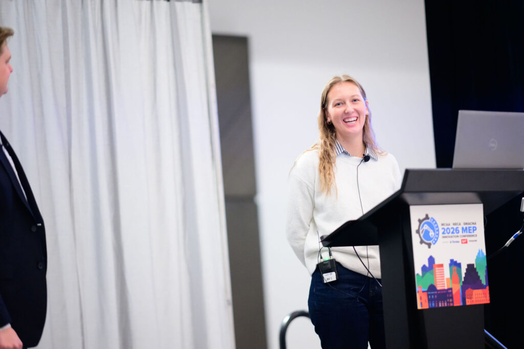 A woman smiles while standing at a podium with a laptop, wearing a microphone headset. A man stands off to the side. The podium sign reads “2026 MEP” and displays illustrated city buildings and gears.