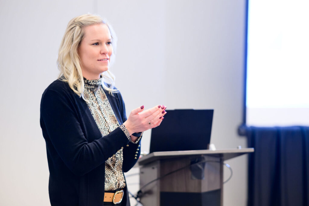 A woman stands next to a podium with a laptop, clapping her hands. She has blonde hair and is wearing a patterned blouse, black cardigan, and beige belt. A projection screen is visible in the background.