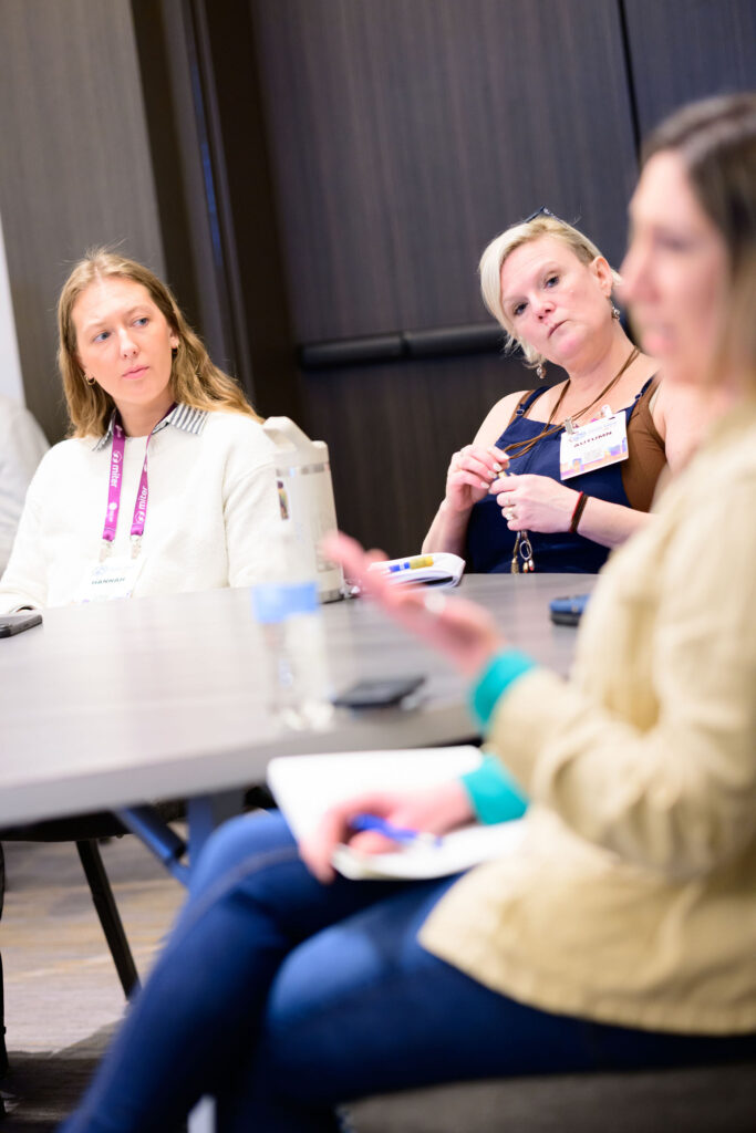 Three women sit around a table in a meeting room, two are attentively listening to the third, who is speaking and holding a notebook. The room has neutral-colored walls and a closed door in the background.