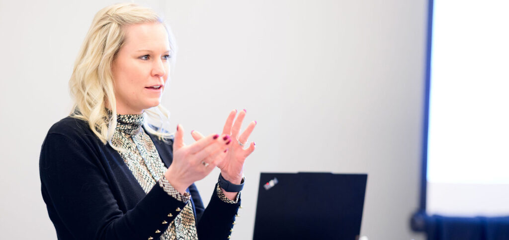 A woman with blonde hair speaks and gestures with her hands while standing behind a laptop in a brightly lit room. She wears a patterned blouse and black cardigan.