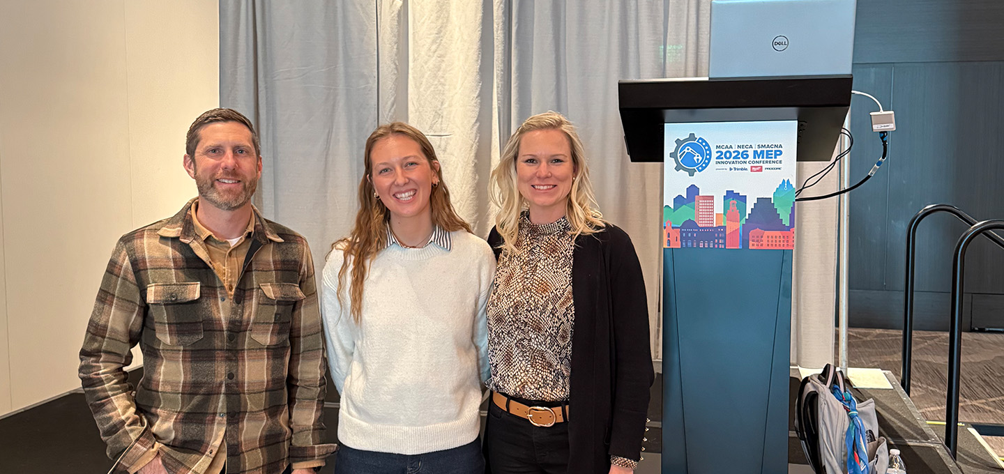 Three people stand indoors in front of a podium with a laptop and a 2026 MEP National Innovation Symposium sign. Curtains and part of a stage are visible in the background.