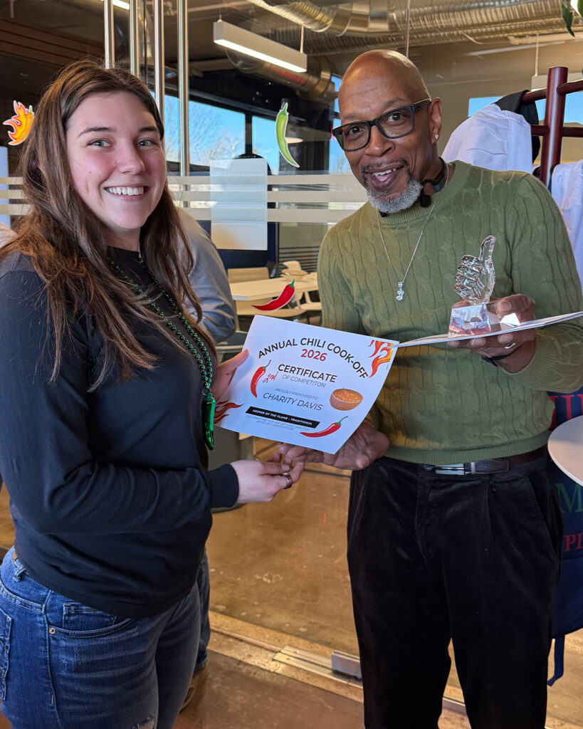 A woman and a man stand indoors. The woman holds a certificate that reads Annual Chili Cook-Off 2024 Certificate, while the man is holding a small trophy. Both are smiling at the camera.