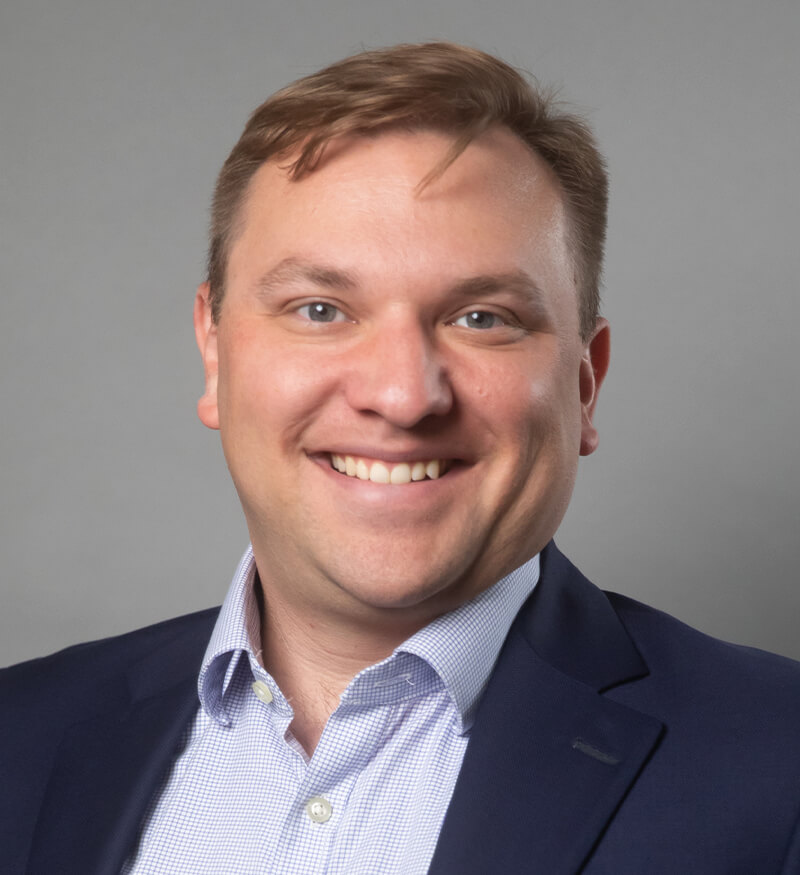 A man with light brown hair, wearing a navy blazer and a light-colored, checked dress shirt, smiles at the camera against a plain gray background.