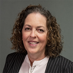 A woman with curly brown hair wearing a black pinstripe blazer over a white blouse is smiling slightly in front of a plain gray background.