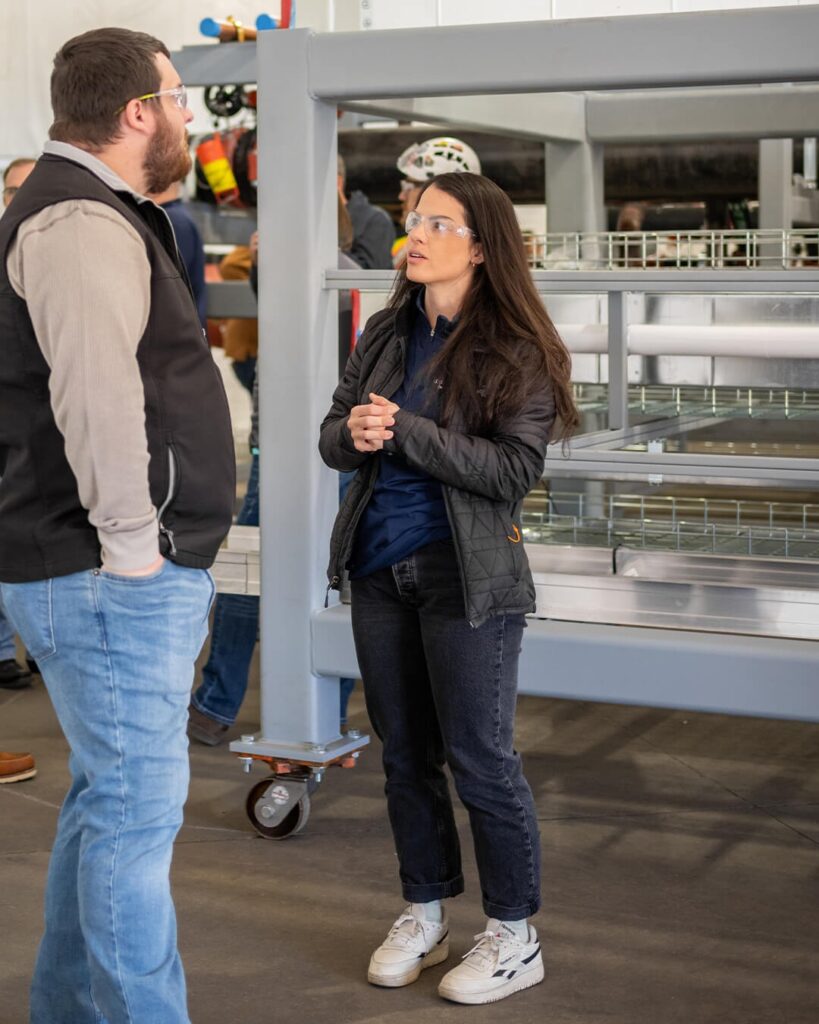 A man and a woman wearing safety glasses are talking in an industrial setting. The woman stands with her hands clasped, and metal racks and equipment are visible in the background.