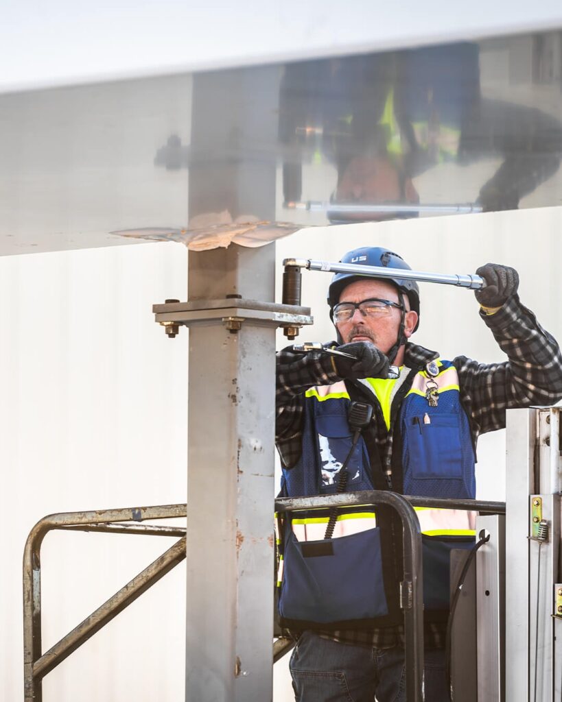 A man wearing safety gear and a blue vest uses a large wrench to tighten a bolt on a metal structure while standing on a raised platform.