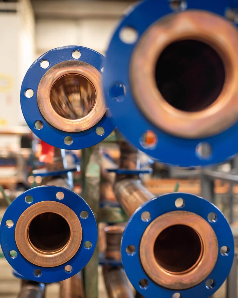 Close-up view of several large metal pipes with blue flanges, arranged horizontally and facing the camera, inside an industrial or factory setting. The background is blurred.
