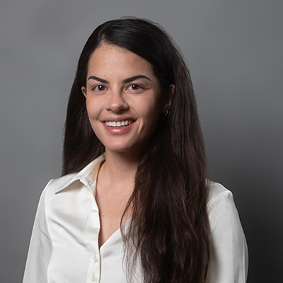 A woman with long dark hair and a white collared shirt smiles at the camera against a plain gray background.