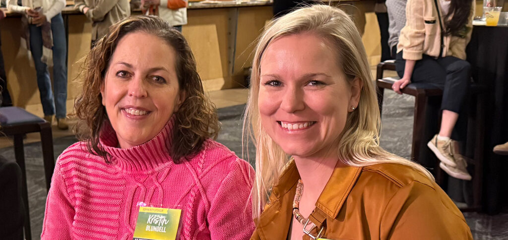 Two women are seated indoors at an event. One woman wears a pink sweater, and the other wears a brown jacket and necklace. People and tables are visible in the background. Both women are smiling at the camera.