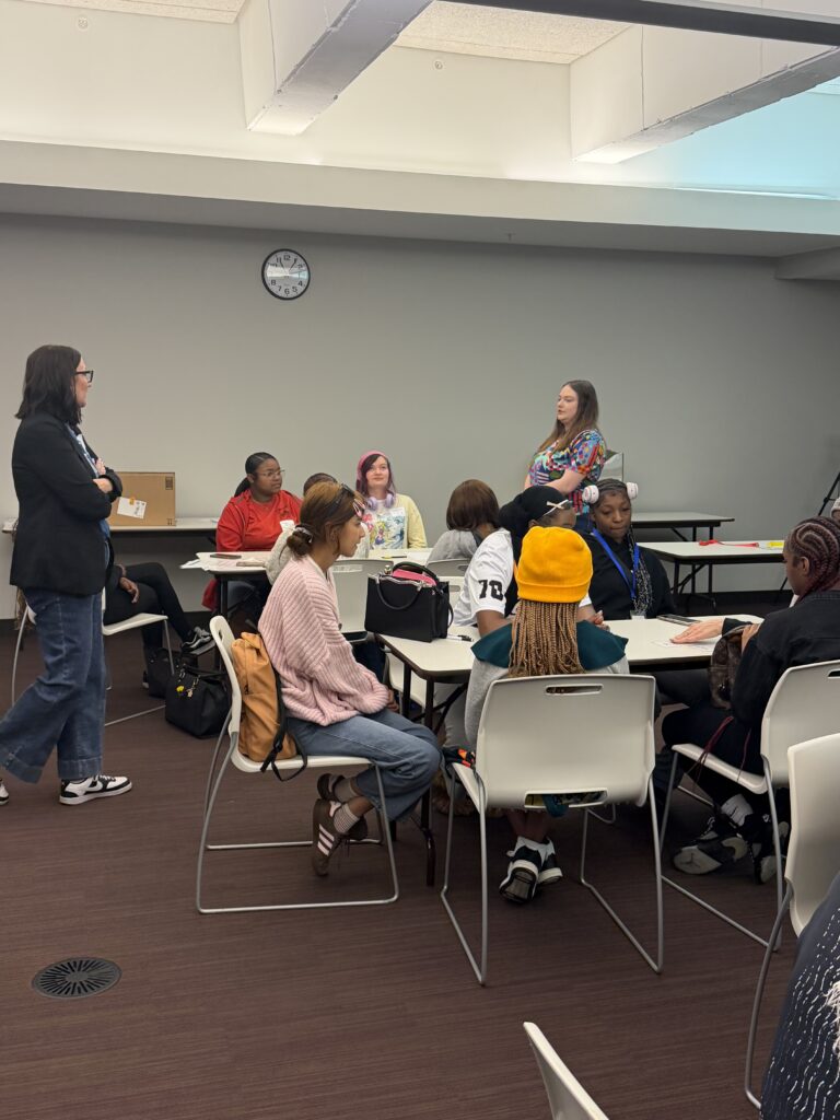 A group of people sit around tables in a meeting room while two women stand and address them. A clock on the wall shows the time as 10:11. The atmosphere appears casual and attentive.