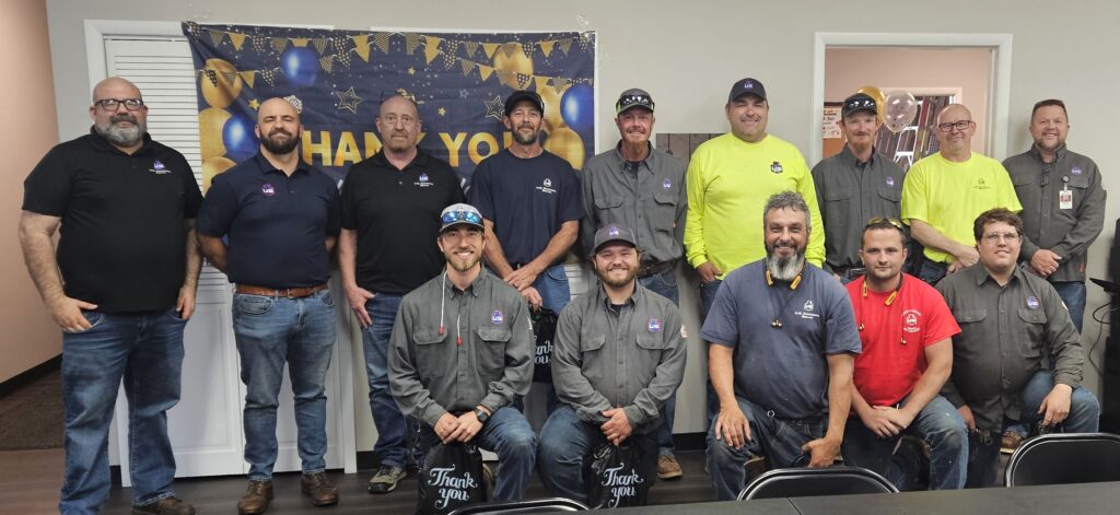 A group of fifteen people, mostly men, pose indoors in front of a Thank You banner. Some wear matching gray uniforms, others wear casual or work clothes in various colors. Several are smiling at the camera.