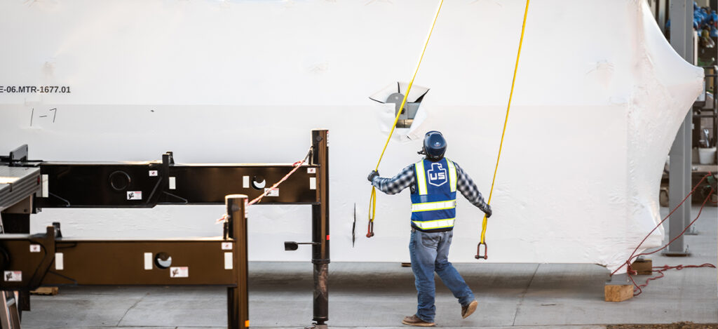 A construction worker in a safety vest and helmet guides two yellow straps attached to a large white steel structure inside an industrial facility.