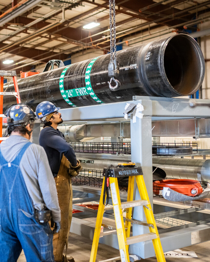 Two workers in protective gear stand near a large black industrial pipe labeled 24 FWR, which is suspended by a chain indoors. A yellow ladder and various equipment are visible in the foreground.
