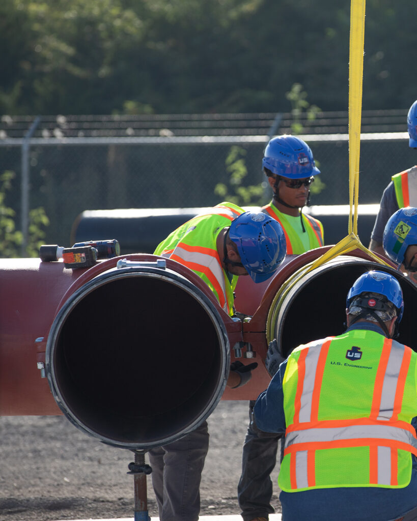 Three workers in safety vests and blue helmets are inspecting or assembling large metal pipes outdoors, using a yellow lifting strap. A fence and trees are visible in the background.