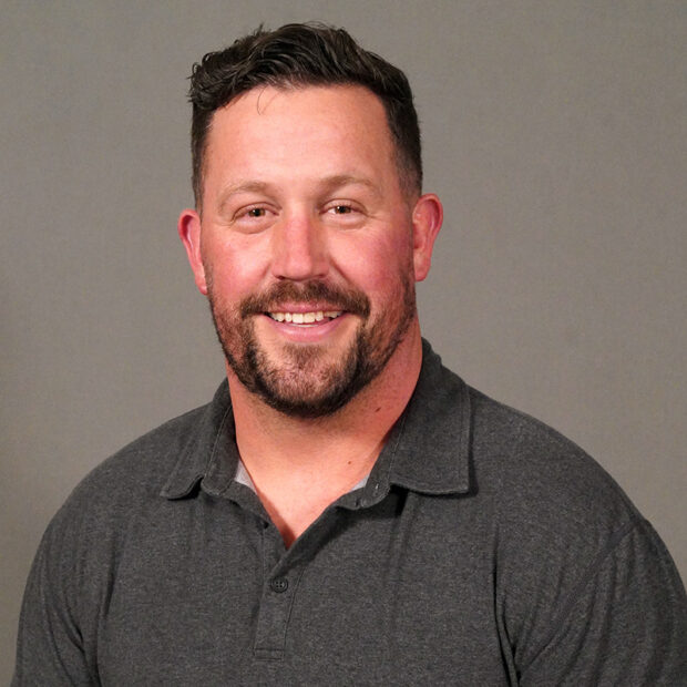 A man with short brown hair and a trimmed beard, wearing a dark gray collared shirt, smiles at the camera in front of a plain gray background.