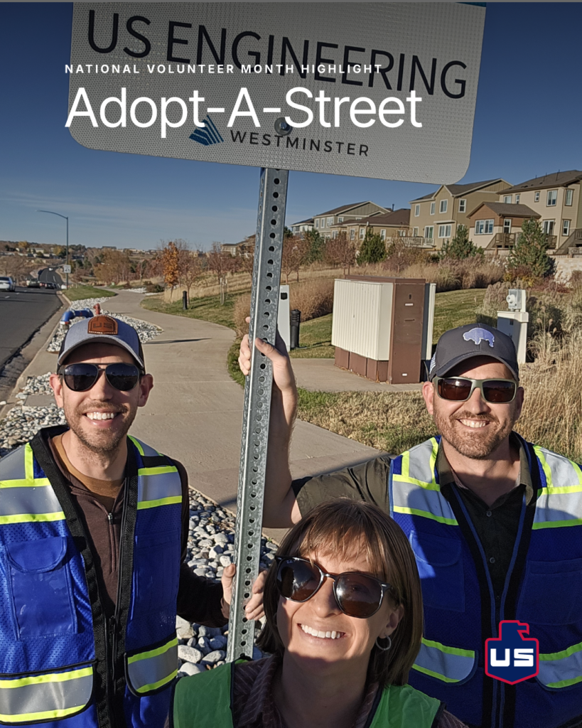 Three adults wearing safety vests and sunglasses stand outdoors on a sidewalk, smiling and holding a sign that reads “US Engineering Adopt-A-Street Westminster.” Houses and trees are visible in the background.