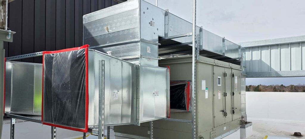 A large commercial HVAC unit with metal ductwork installed on a rooftop. Some duct sections are covered with protective plastic sheets. The background shows part of the building structure and an overcast sky.