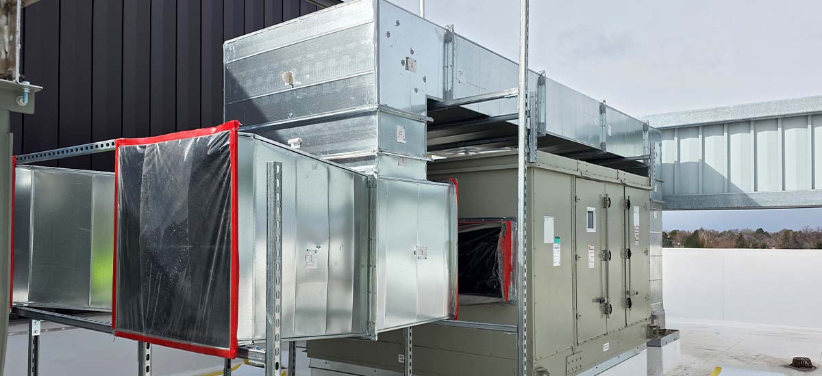 A large commercial HVAC unit with metal ductwork installed on a rooftop. Some duct sections are covered with protective plastic sheets. The background shows part of the building structure and an overcast sky.