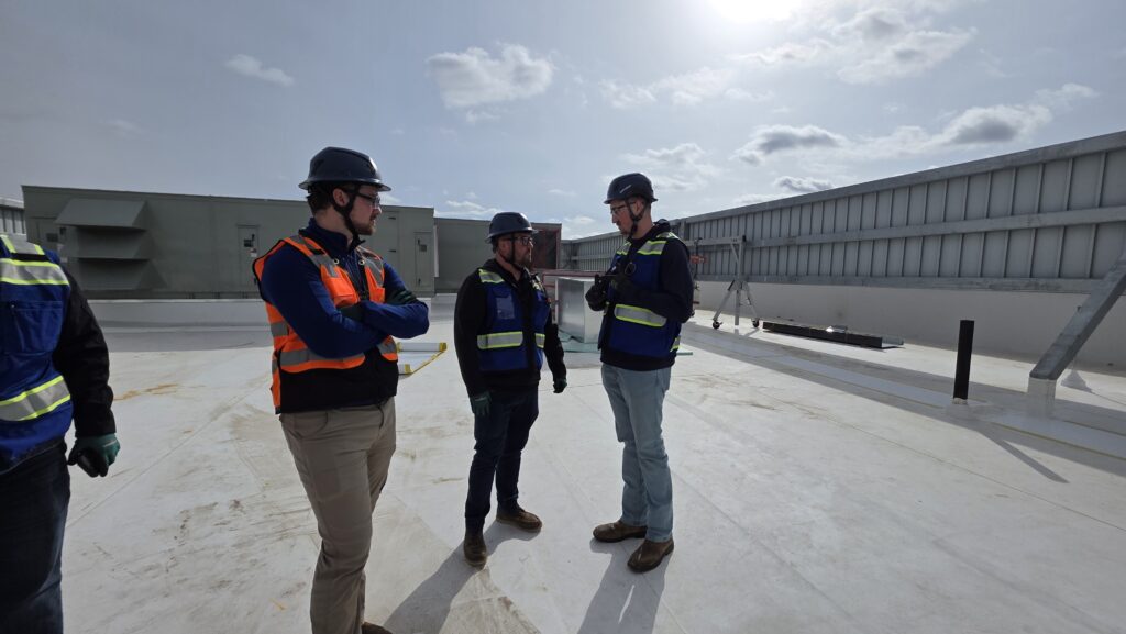 Three people wearing safety vests, helmets, and protective gear stand on a flat rooftop in daylight, having a discussion. Industrial equipment and railings are visible in the background under a partly cloudy sky.