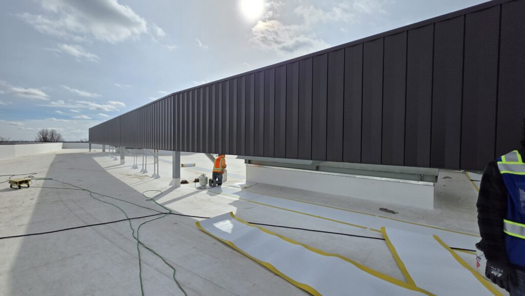 Workers in safety vests install or inspect equipment under a large, dark panel on a flat rooftop. White material and tools are spread on the ground, with the sky partly cloudy and the sun visible.