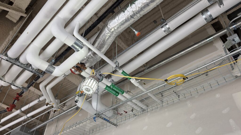 Ceiling view of exposed white utility pipes, insulated ducts, wiring, and light fixtures in an unfinished building interior, with a cable tray and wired emergency light visible.