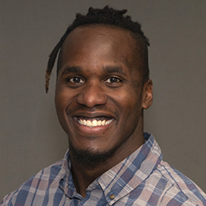 A man with short hair and a small braid smiles at the camera. He is wearing a blue and gray plaid button-up shirt and is posed against a plain gray background.