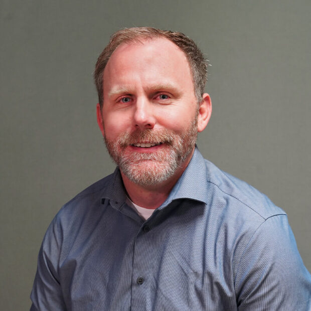 A middle-aged man with short light brown hair and a beard is wearing a blue button-up shirt. He is smiling and seated in front of a plain gray background.