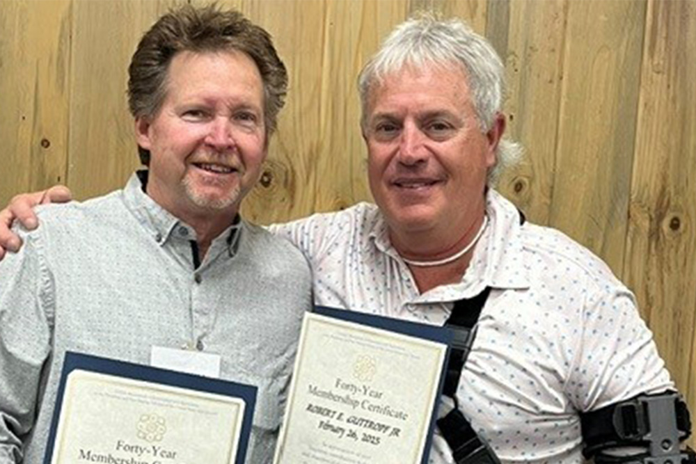 Two men stand side by side in front of a wooden wall, each holding a certificate titled Forty-Year Membership Certificate. One man has his arm around the other and wears a shoulder brace. Both are smiling at the camera.