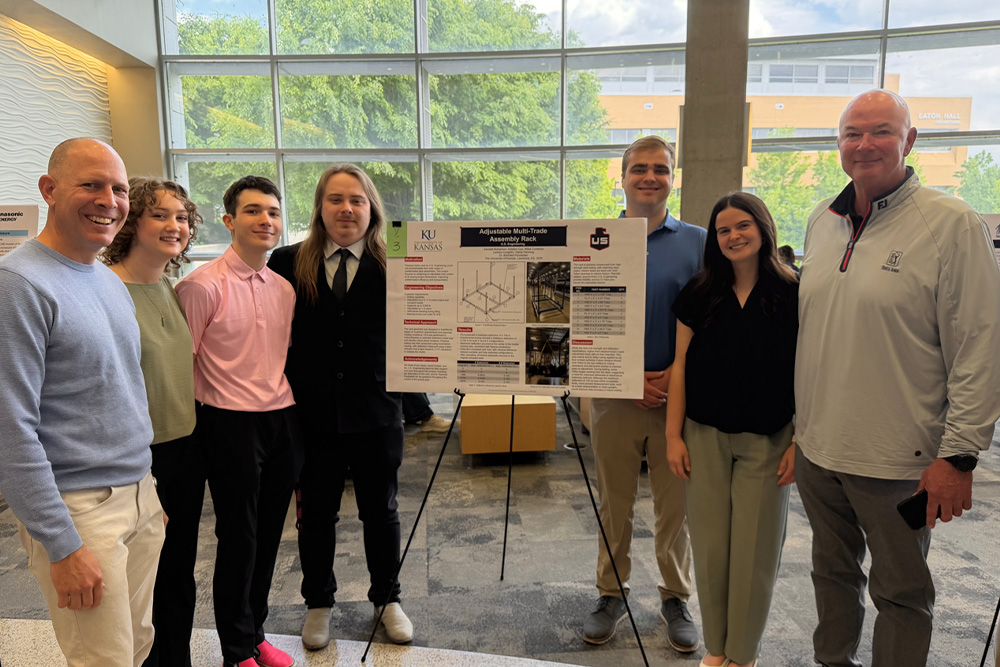 Seven people stand indoors in front of a large window, posing around a poster displayed on an easel. The poster appears to be an academic or project presentation. Everyone is smiling at the camera.