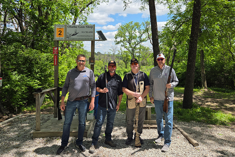 Four men stand outdoors at a shooting range under a sign reading “Olathe Gun Shop.” They are smiling, holding firearms, and surrounded by trees and gravel on a sunny day.