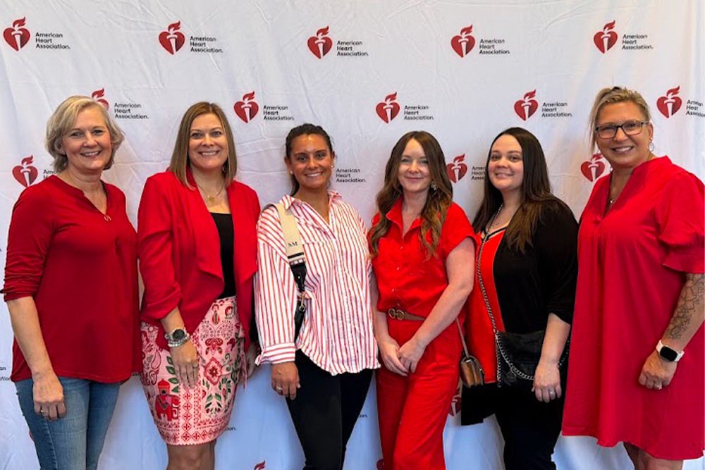 Six women stand in front of an American Heart Association backdrop, most wearing red clothing. They are posing together and smiling for the photo.