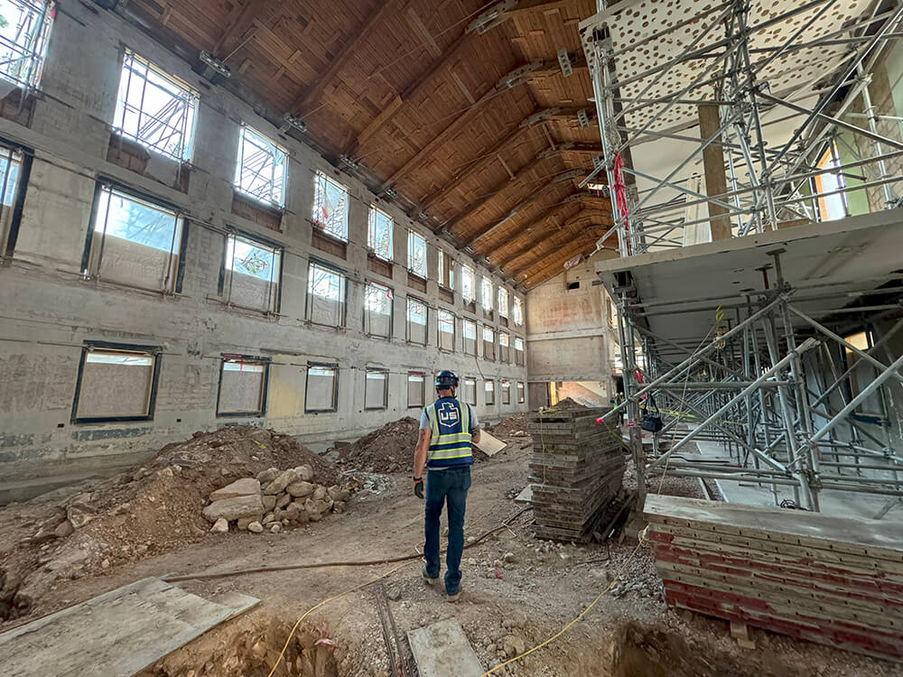 A construction worker wearing a safety vest and helmet stands inside a large, partially renovated building with exposed brick, scaffolding, dirt piles, and unfinished walls. Natural light enters through multiple windows.