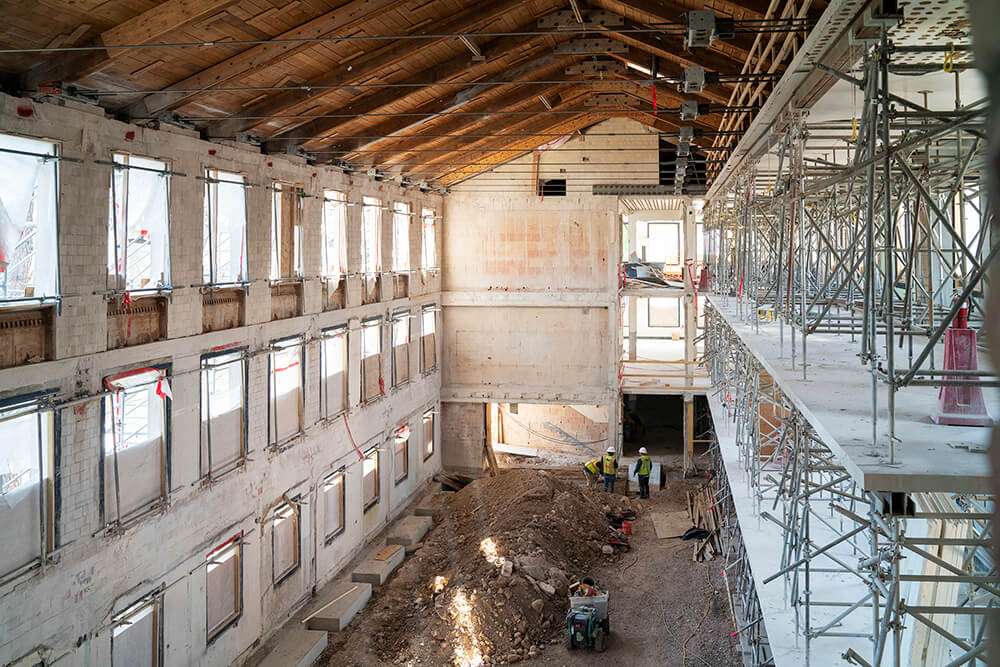 A large, partially renovated building interior with exposed wooden beams, scaffolding on the right, piles of dirt and construction equipment on the ground, and workers gathered near the back wall.