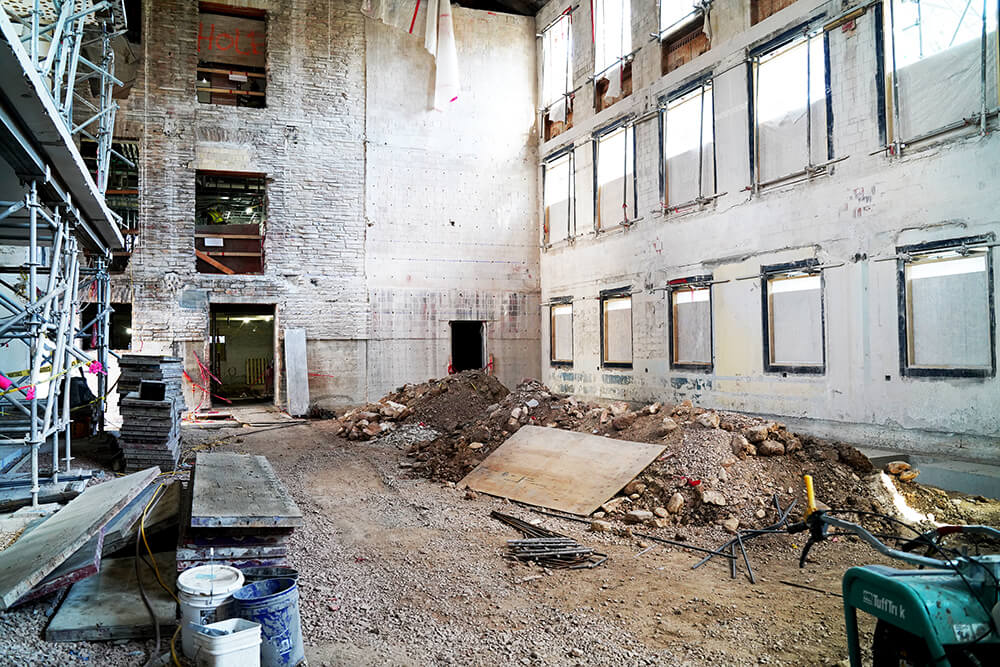 An interior construction site with exposed brick walls, scaffolding on the left, piles of dirt and rubble in the center, covered windows, construction materials, and tools scattered on the ground.