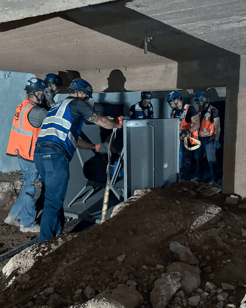 Several construction workers wearing safety gear and helmets are moving a large metal unit under a concrete structure, surrounded by dirt and construction materials.