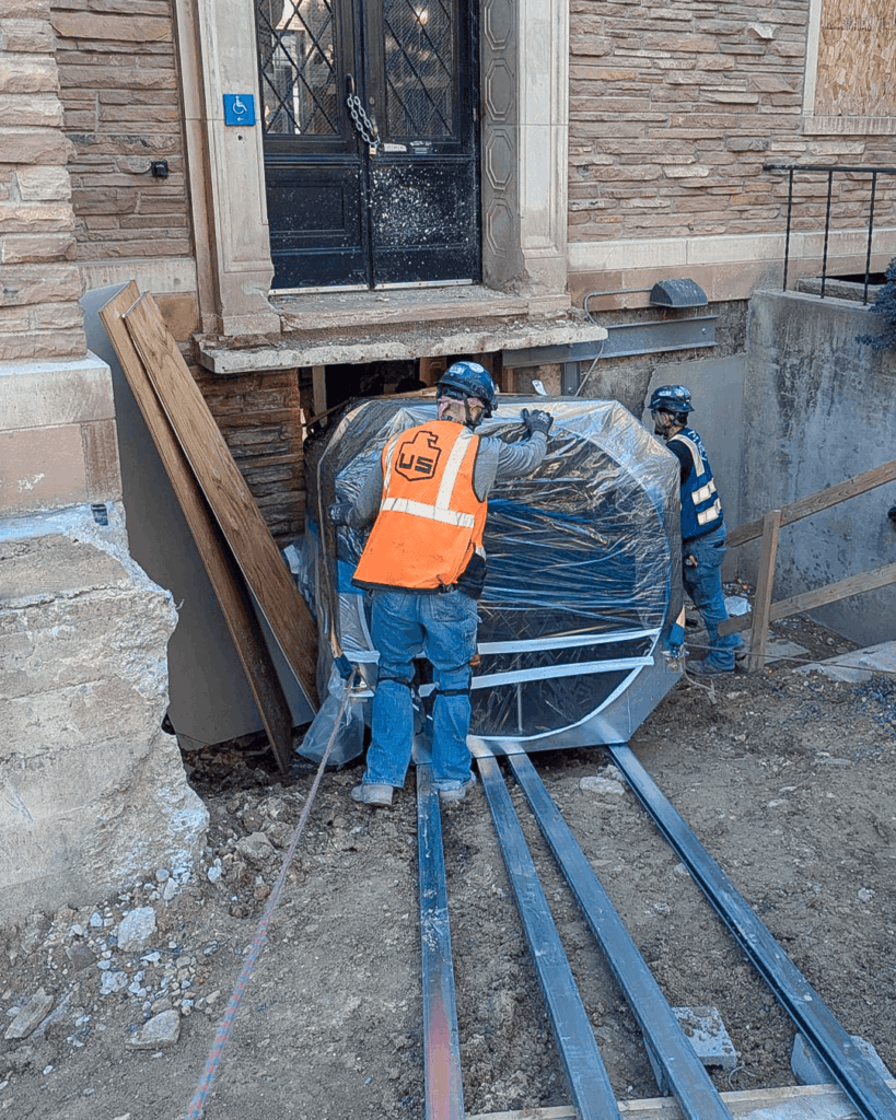 Two construction workers guide a large, wrapped object up metal rails toward a basement entrance of a stone building. One worker wears an orange safety vest; both wear hard hats and safety gear.