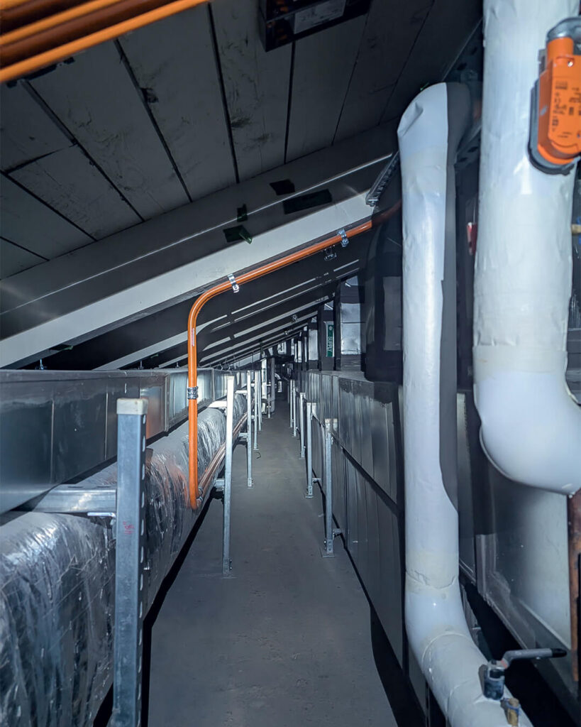 View of a narrow attic space with exposed ductwork, pipes wrapped in insulation, metal supports, and electrical conduits running along the sloped ceiling. The area is illuminated by artificial light.