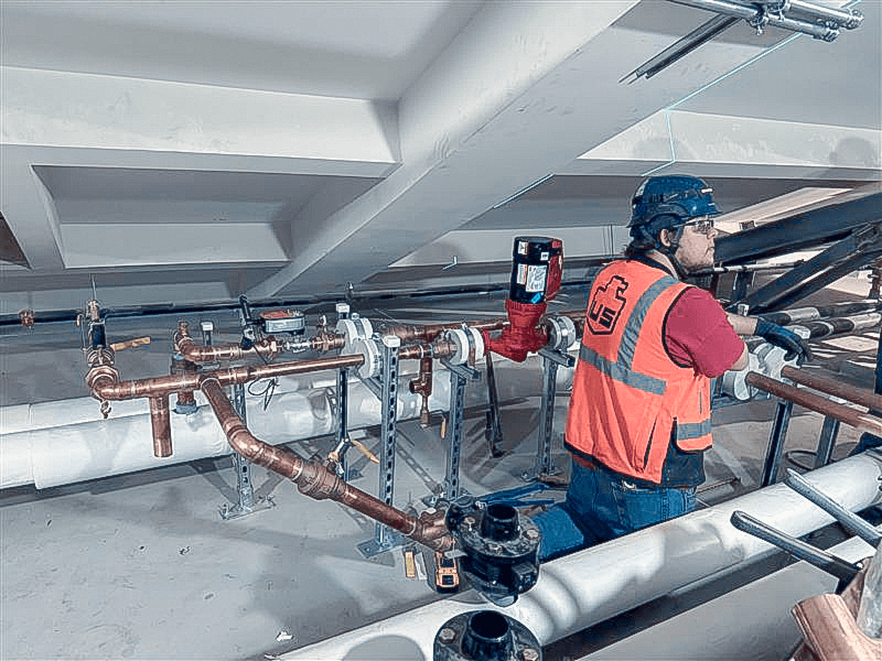 A construction worker wearing a hard hat and orange safety vest inspects and works on an array of copper pipes and valves mounted to supports beneath a building ceiling.