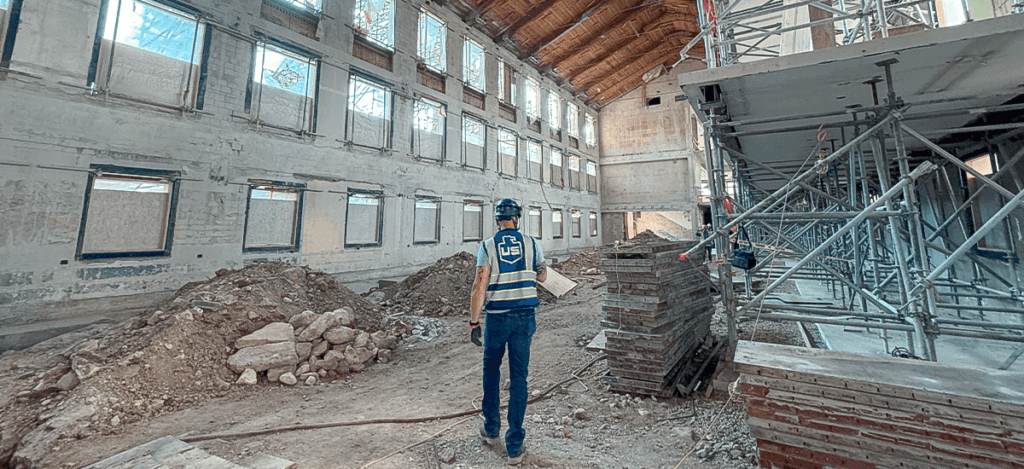 A person wearing a safety vest and helmet stands in a large, unfinished building with exposed walls, piles of dirt and rubble, and scaffolding on the right side. Light enters through tall windows without glass.