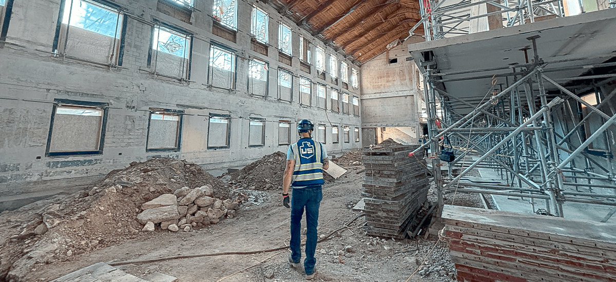 A person wearing a safety vest and helmet stands in a large, unfinished building with exposed walls, piles of dirt and rubble, and scaffolding on the right side. Light enters through tall windows without glass.
