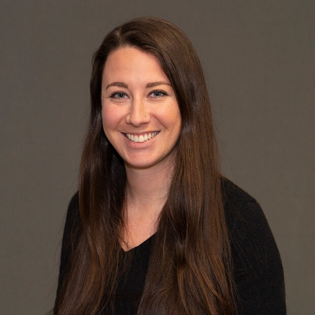 A woman with long brown hair and a black top smiles at the camera in front of a plain gray background.