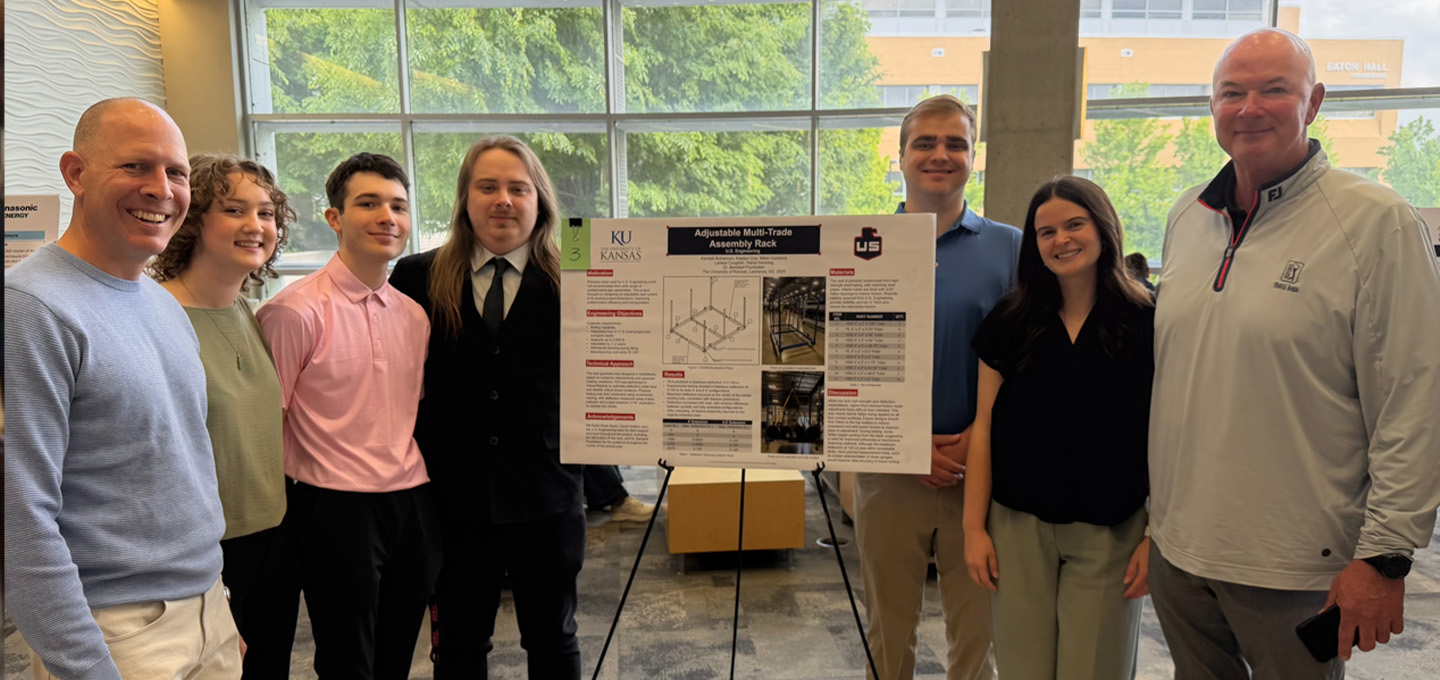 Six people stand indoors in front of a window and smile at the camera. They are gathered around a presentation board titled “Adjustable Hub Truck Assembly Rack,” featuring diagrams and text.