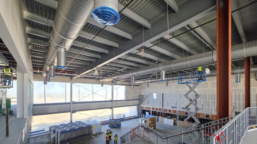 Large industrial interior under construction with exposed ductwork, steel beams, and partially finished walls. Several workers in safety vests are on the ground and one is elevated on a scissor lift. Building materials are scattered around.