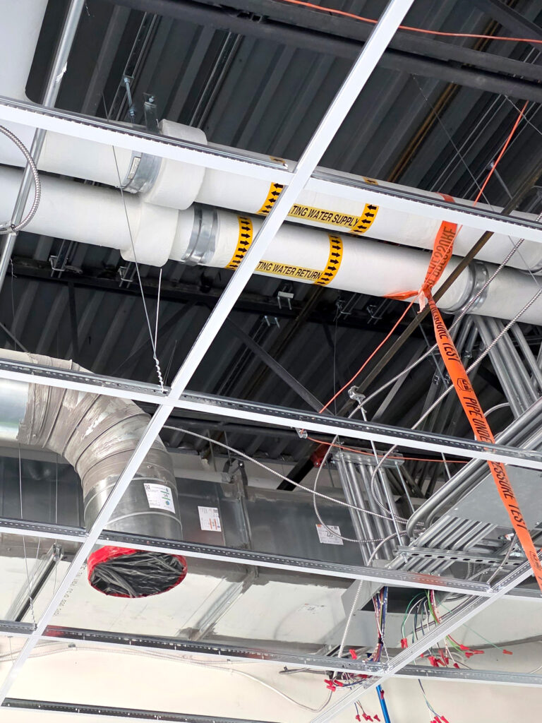 Exposed pipes and ductwork above a drop ceiling grid in a building under construction. Yellow caution tape labels some pipes, and loose wires hang down. The ceiling tiles have not been installed.