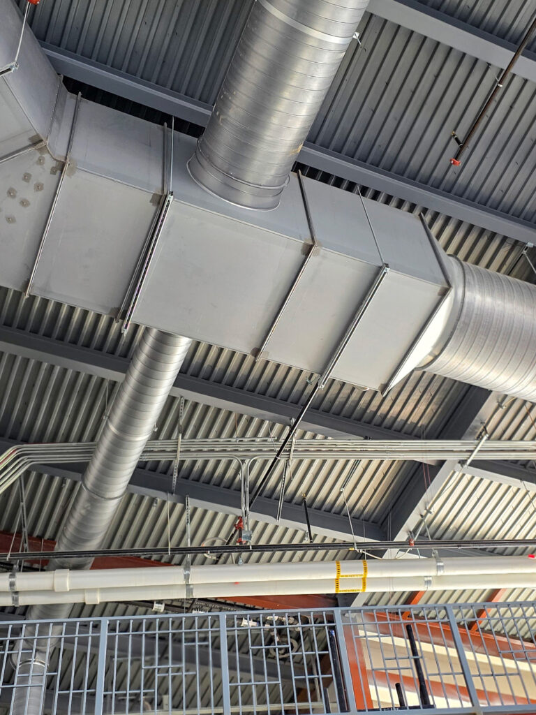 Large metal air ducts and pipes run along a corrugated metal ceiling in an industrial or commercial building. Electrical conduits and other utility lines are also visible.