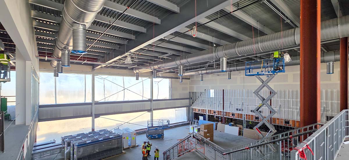 A large indoor construction site with exposed ceiling ducts, unfinished walls, and multiple workers in safety vests. One worker is on a scissor lift, others are gathered on the ground. Building materials are stacked nearby.