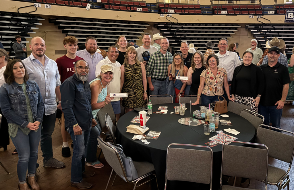 A group of about 20 adults and teens stand and smile around a round table covered with programs, cups, and plates in an indoor arena with empty bleachers in the background.