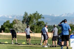 A group of people are practicing golf at a driving range on a sunny day, with golf clubs and bags nearby and mountains visible in the background.