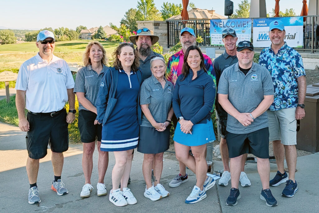 A group of ten adults pose together outdoors on a golf course. Most are wearing golf shirts and shorts or skirts, with one person in a colorful tie-dye shirt. A Welcome sign is visible in the background.