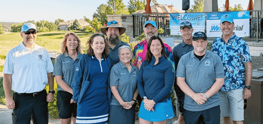 A group of ten people, wearing casual and golf attire, stand outdoors smiling at the camera. Some wear matching gray shirts with a logo. There is a golf course and welcome banners visible in the background.